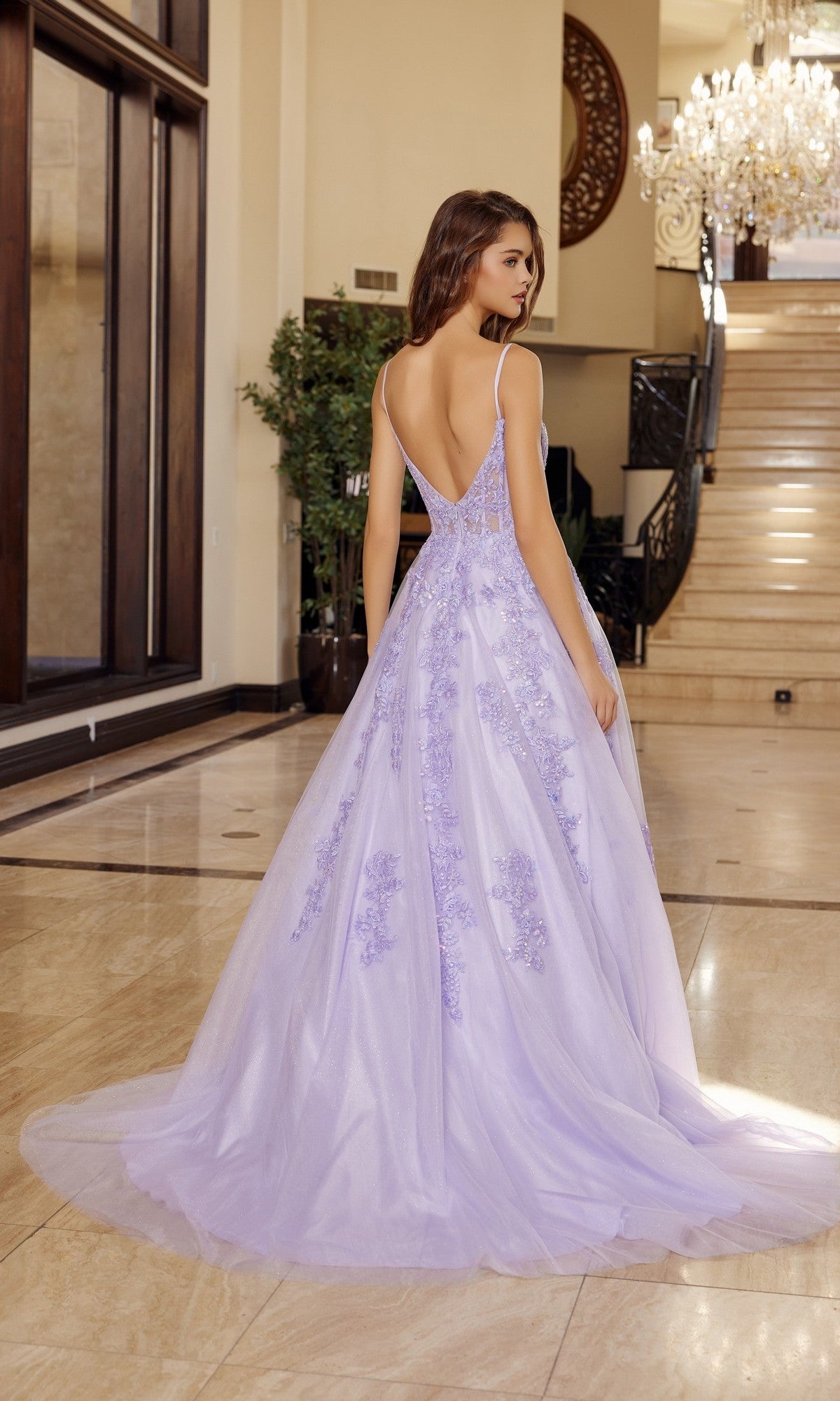 A woman stands indoors on a marble floor, facing away from the camera, wearing the Nox Anabel H1880 long prom dress—a floor-length lilac gown with a sequin corset bodice and floral embellishments.